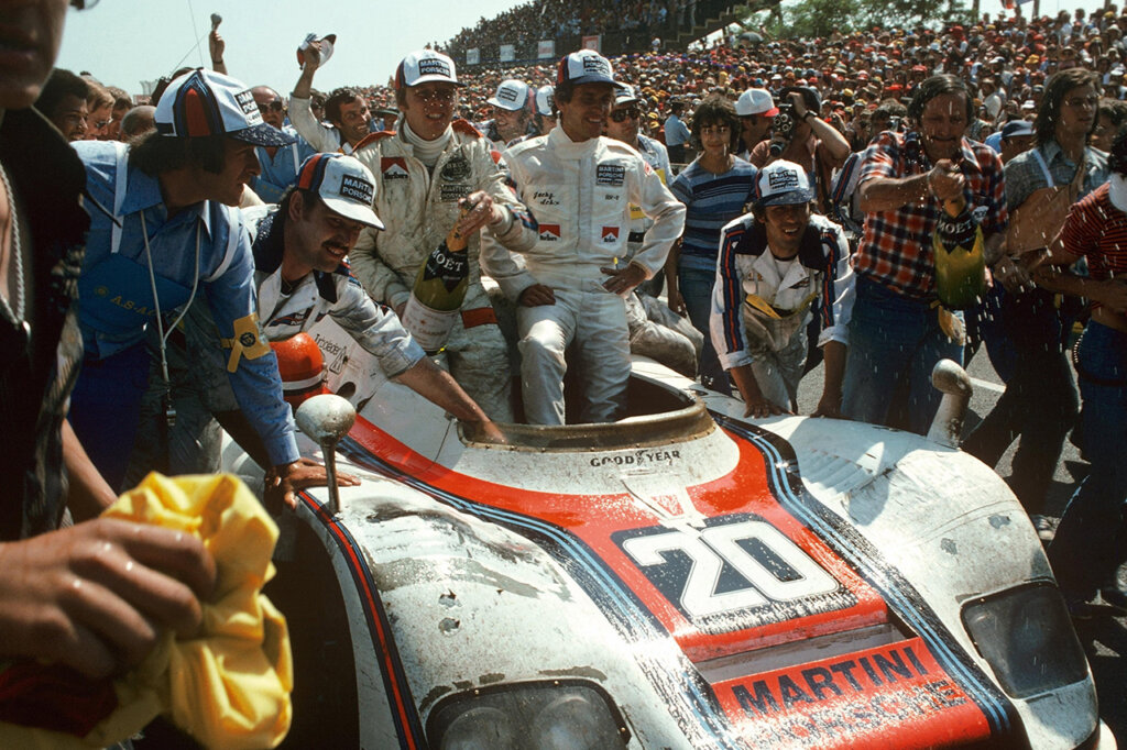 Two men in white race suits sitting in a Porsche 936 race car with Martini race livery surrounded by many people celebrating with them.