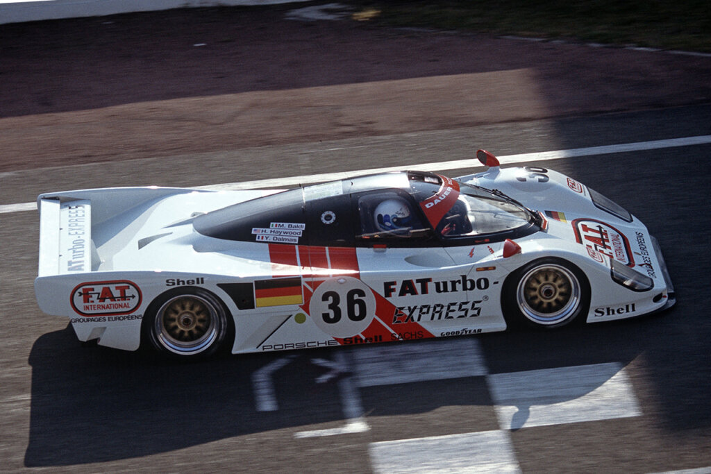 White Porsche 962 Dauer Le Mans GT race car with orange and red accent striping crossing a finish line on a racetrack.