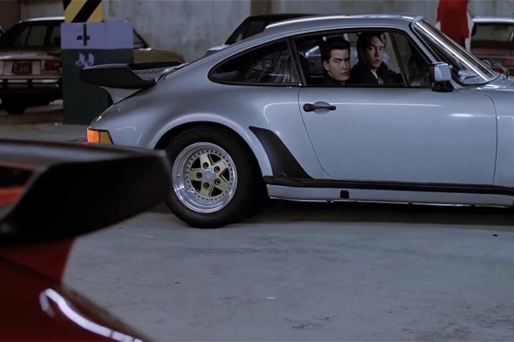Charlie Sheen sitting in the passenger seat of a silver Porsche 911 930 Turbo looking at a Red 911 inside of a parking garage.