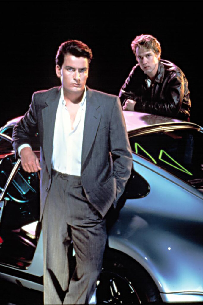 Two men standing next to a Silver Porsche one on each side of the car with a black background.