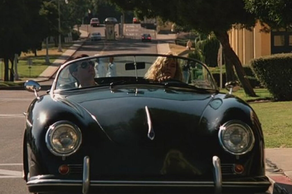 Black Porsche 356A driven by a lady with blonde hair and actor Tom Cruise sitting in the passenger seat with homes, green grass and road in background.