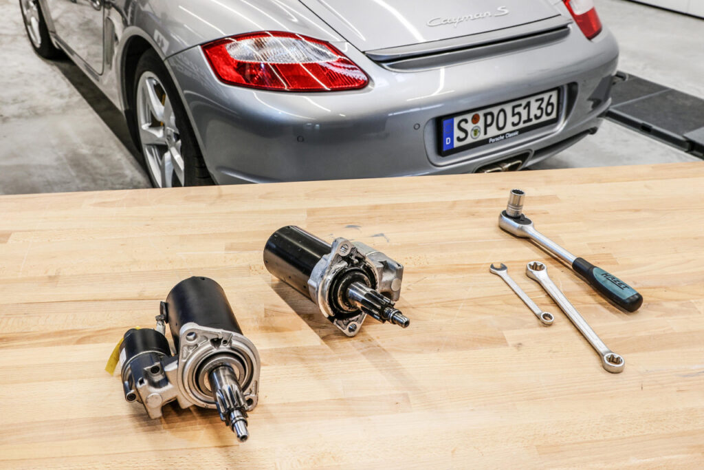 Porsche parts and tools on a table with a rear of a silver porsche cayman s in the background