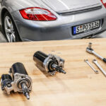 Porsche parts and tools on a table with a rear of a silver porsche cayman s in the background